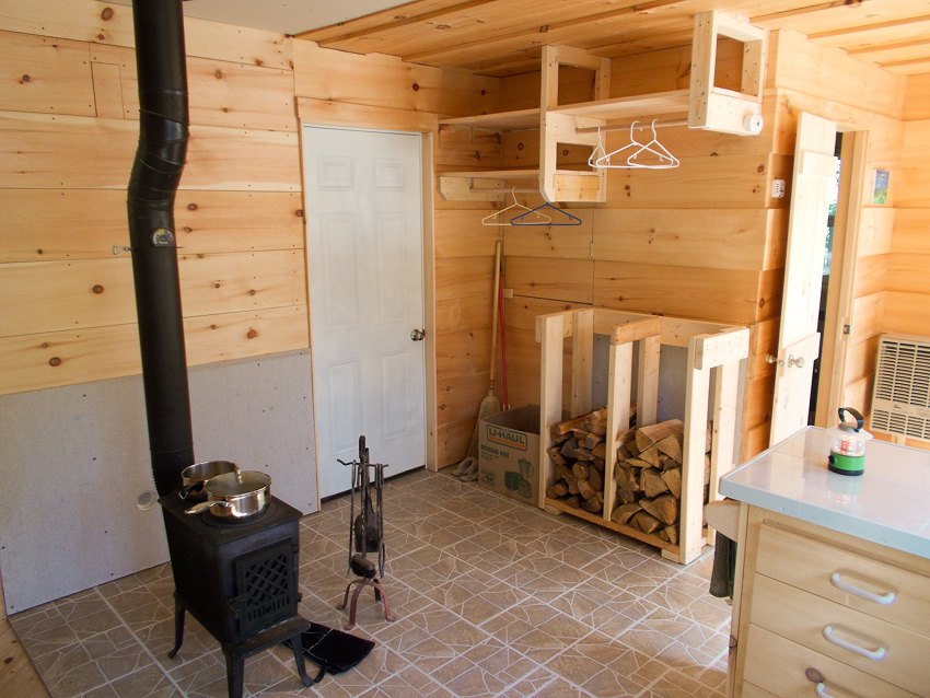 Woodstove faces into the room beside the front door. On the other side of the door are a firewood storage rack, hanging space, and shelves